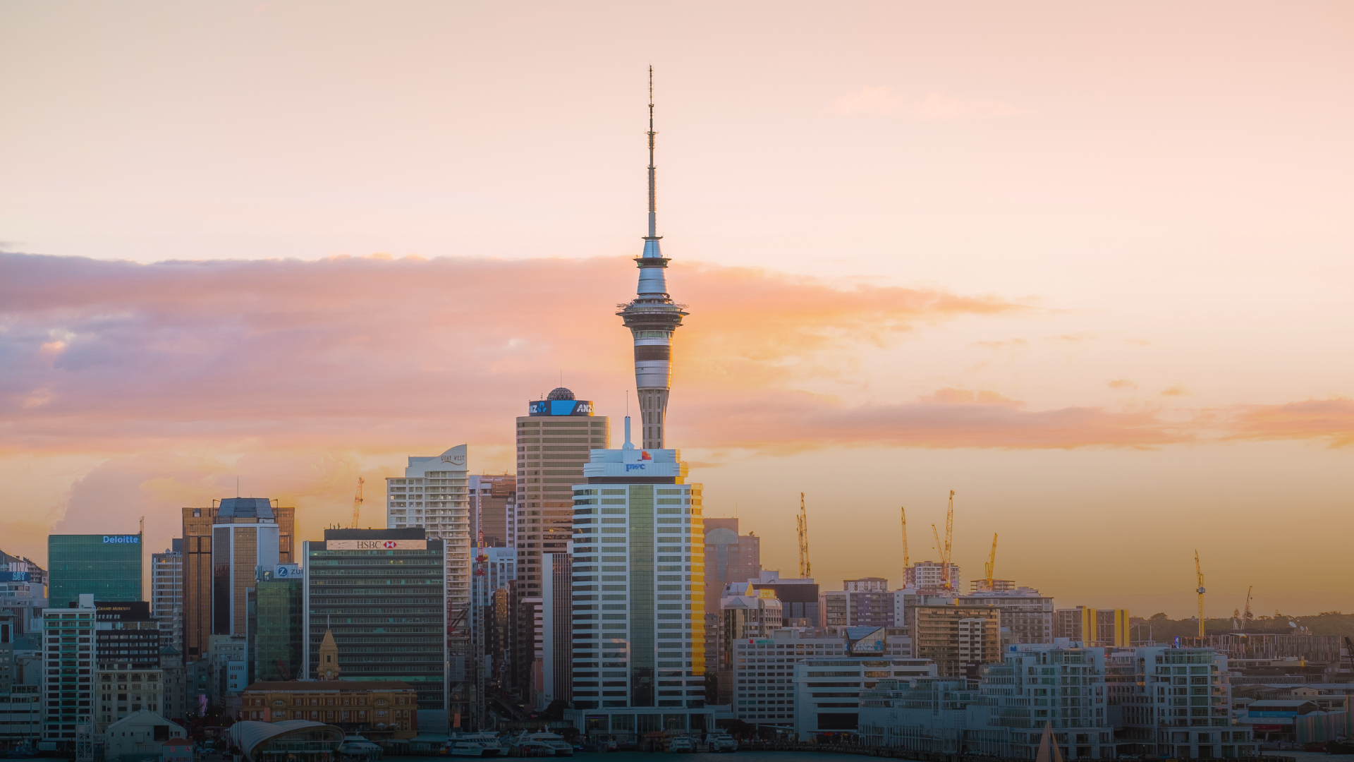 Sky Tower and Auckland skyline at sunset. Orange and pink sky, modern buildings.