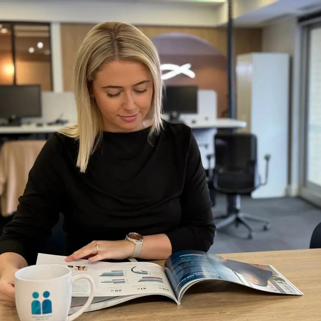 Woman with blonde hair looks at a catalog at a desk in an office. She wears a black top, has a watch and a mug.