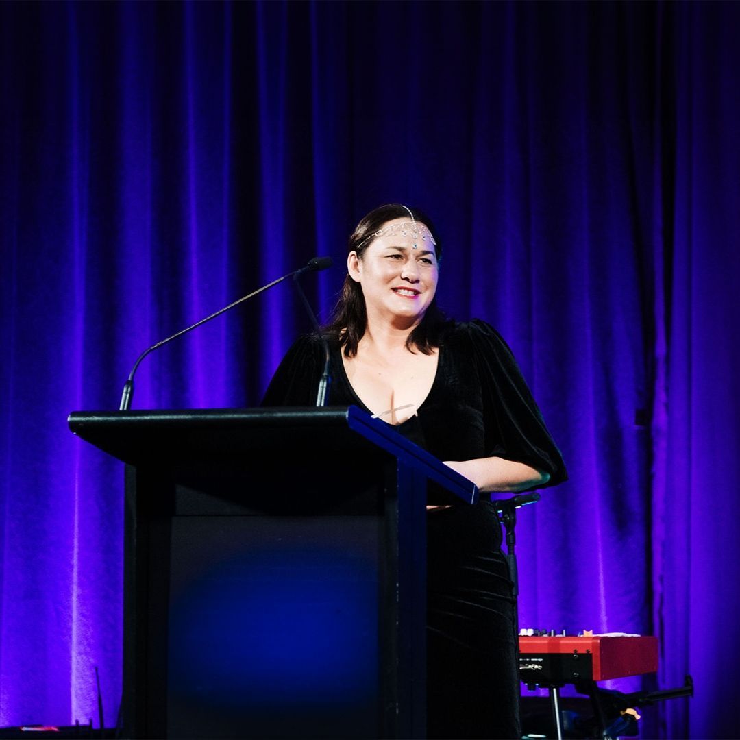 Woman speaking at a podium, smiling, with blue curtain backdrop.