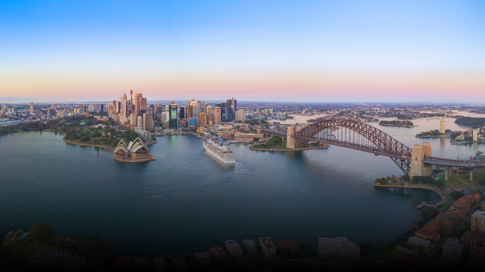 Sydney Harbour with city skyline, Opera House, Harbour Bridge, and cruise ships at dawn.