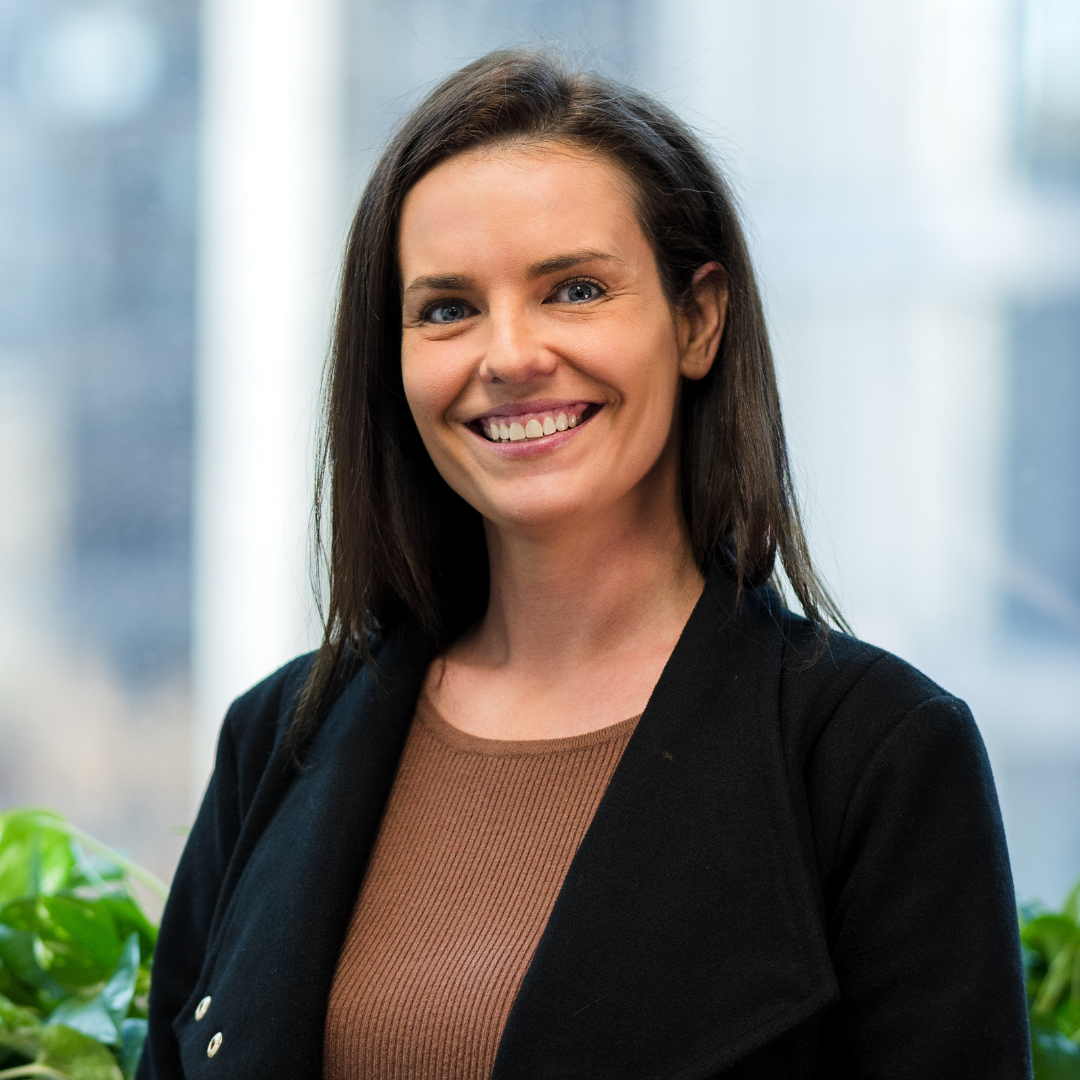Woman in black jacket smiles, brown shirt, city background.