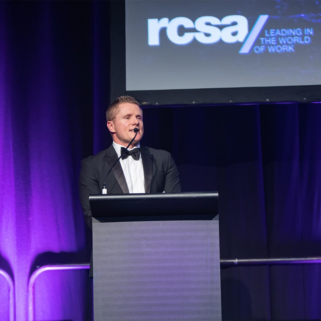 Man in tuxedo at podium, speaking. RCSA logo on screen in background. Purple lighting.