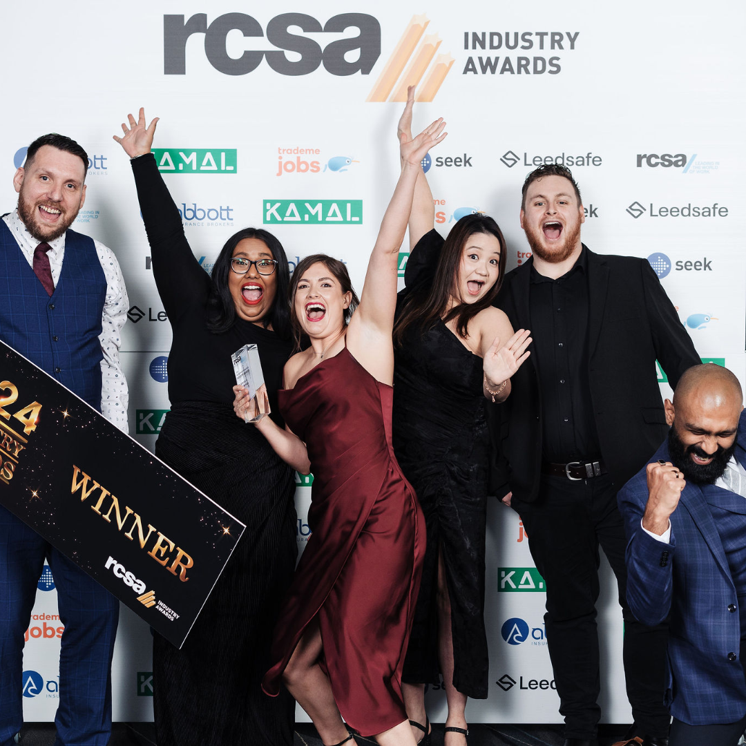 A group celebrates a win at an industry awards event. People are raising arms, smiling, and holding a winner's check.