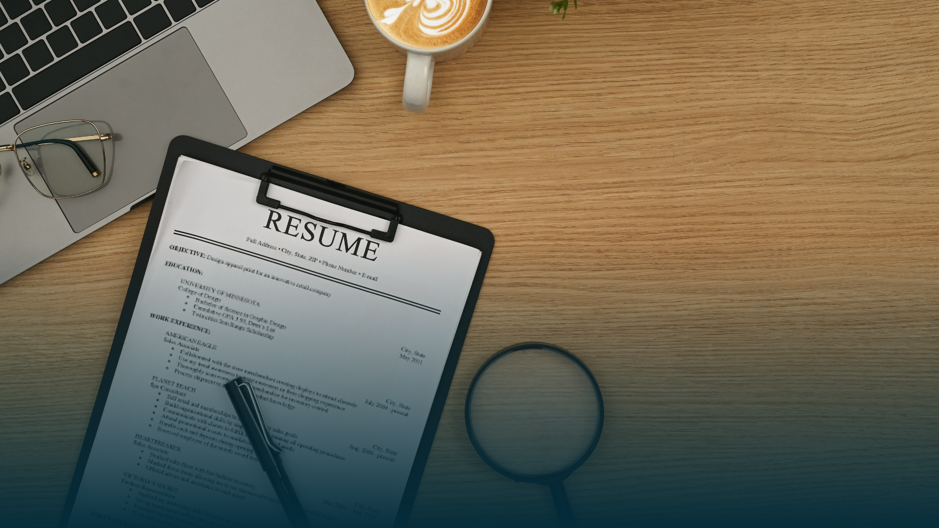 Resume on clipboard with laptop, glasses, magnifying glass, pen, and coffee on a wooden desk.