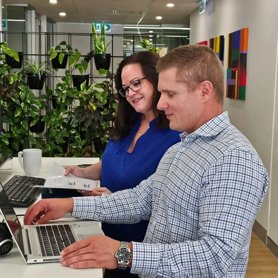 Two people reviewing paperwork and using a laptop in an office. Plants and artwork in background.