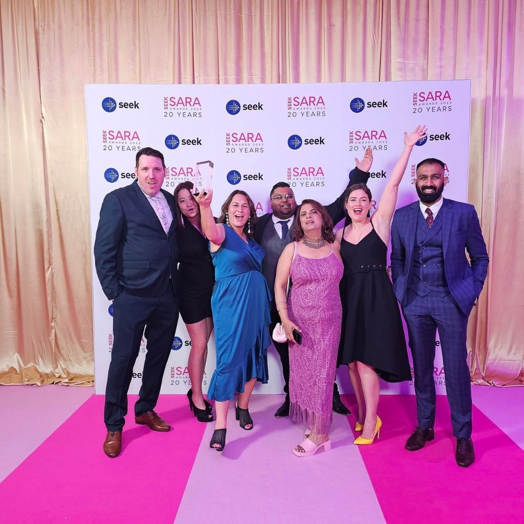 Group of people celebrating, holding award on pink carpet; backdrop with logos.