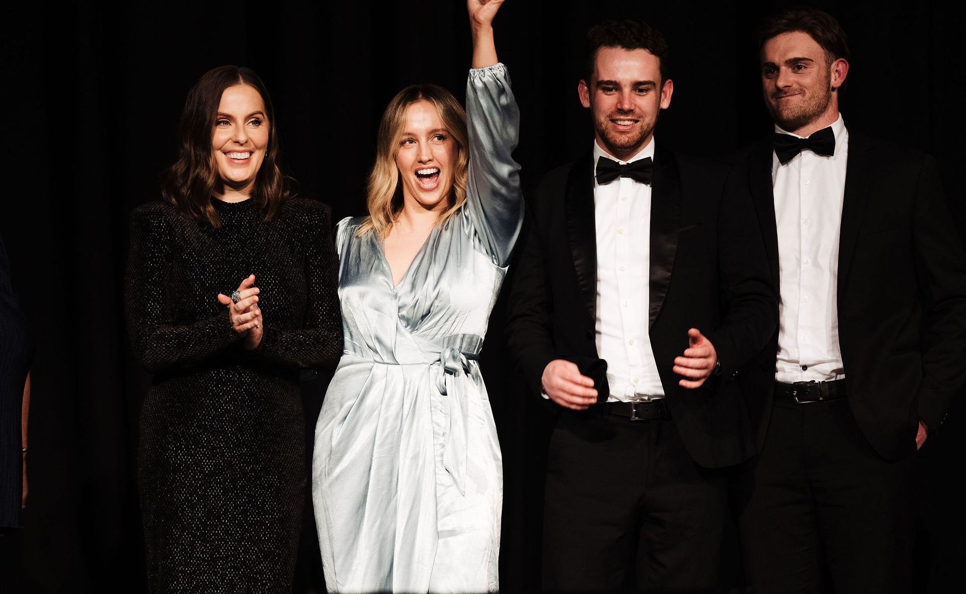 Four people on stage, one raising arm. Woman in blue dress celebrates. Others in formal wear applaud.