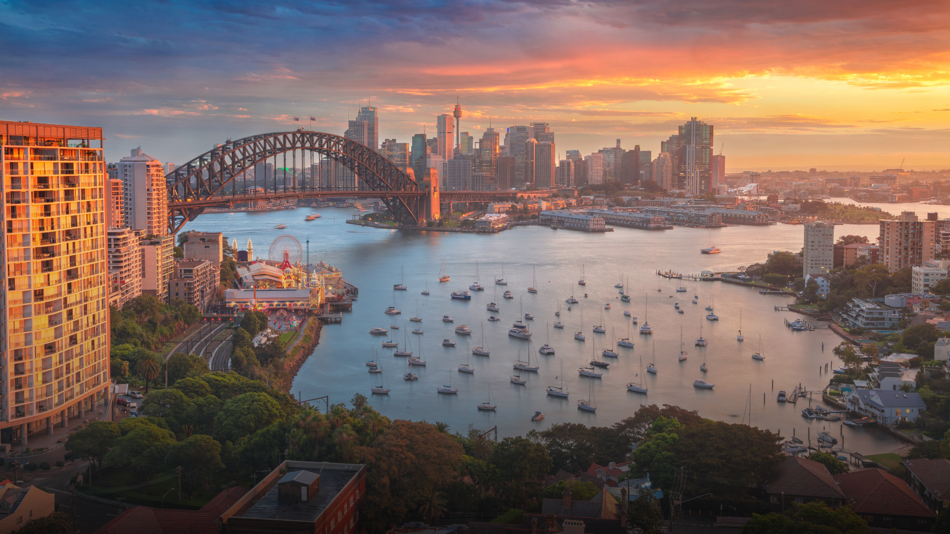 Sydney Harbour at sunset with Harbour Bridge, cityscape, and boats. Orange and blue sky.