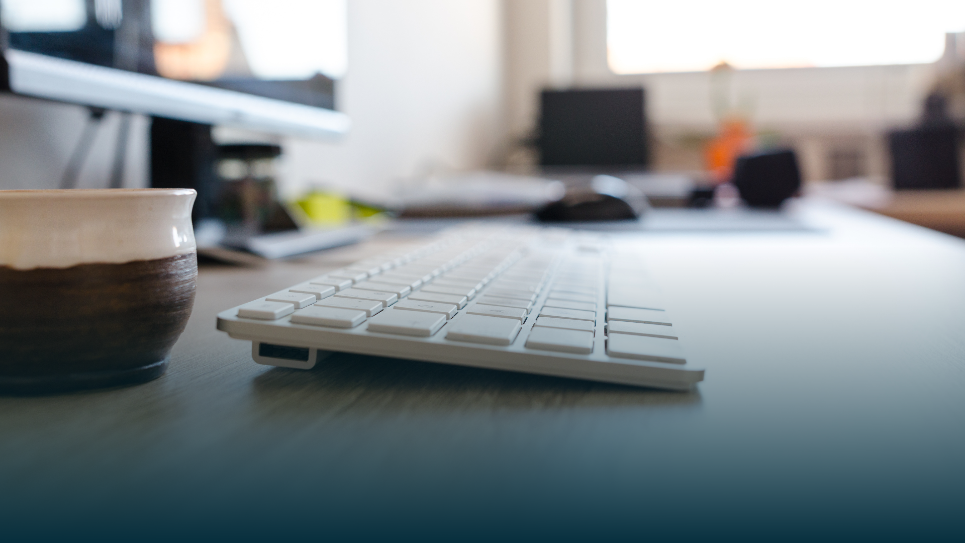 Keyboard and coffee mug on desk in office setting; computer in background.