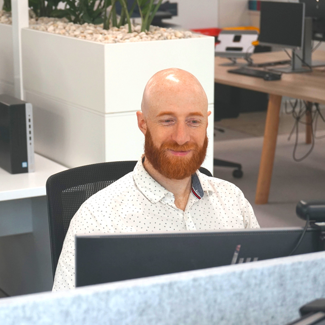 Man with red beard smiles at a computer in a modern office.