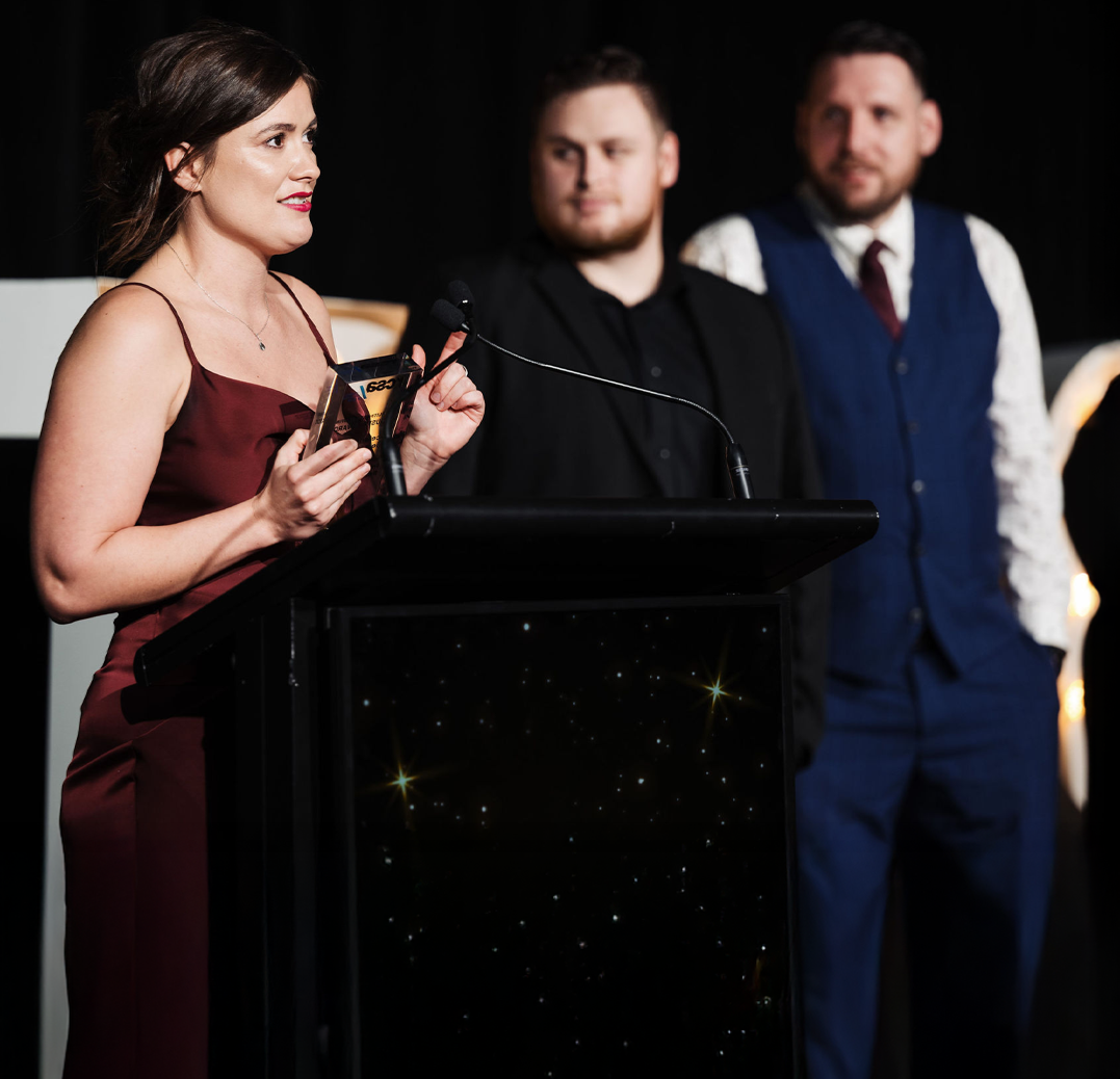 Woman in burgundy dress speaking at a podium, holding award; two men behind her.