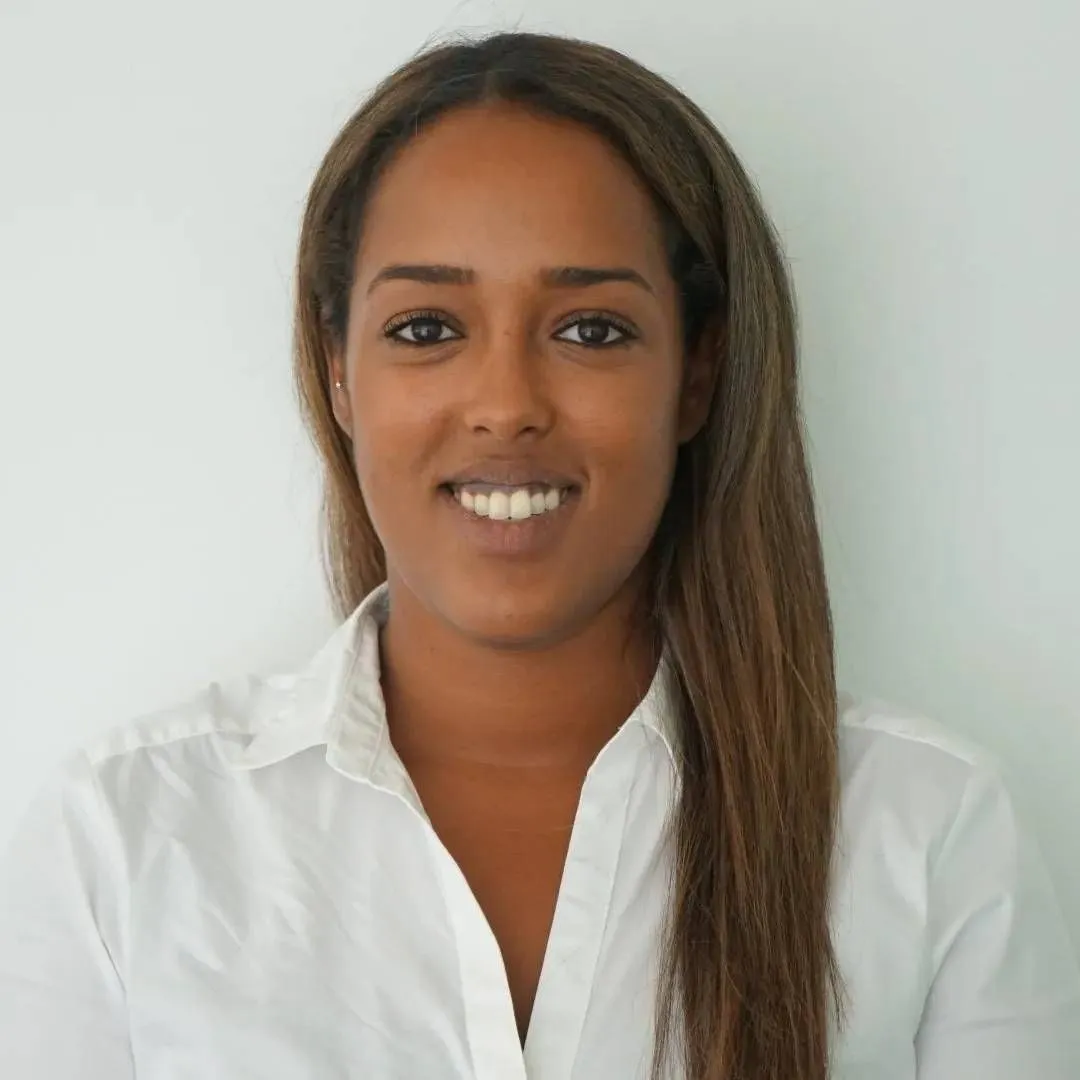 Woman with long brown hair smiling, wearing a white button-down shirt, in front of a white background.