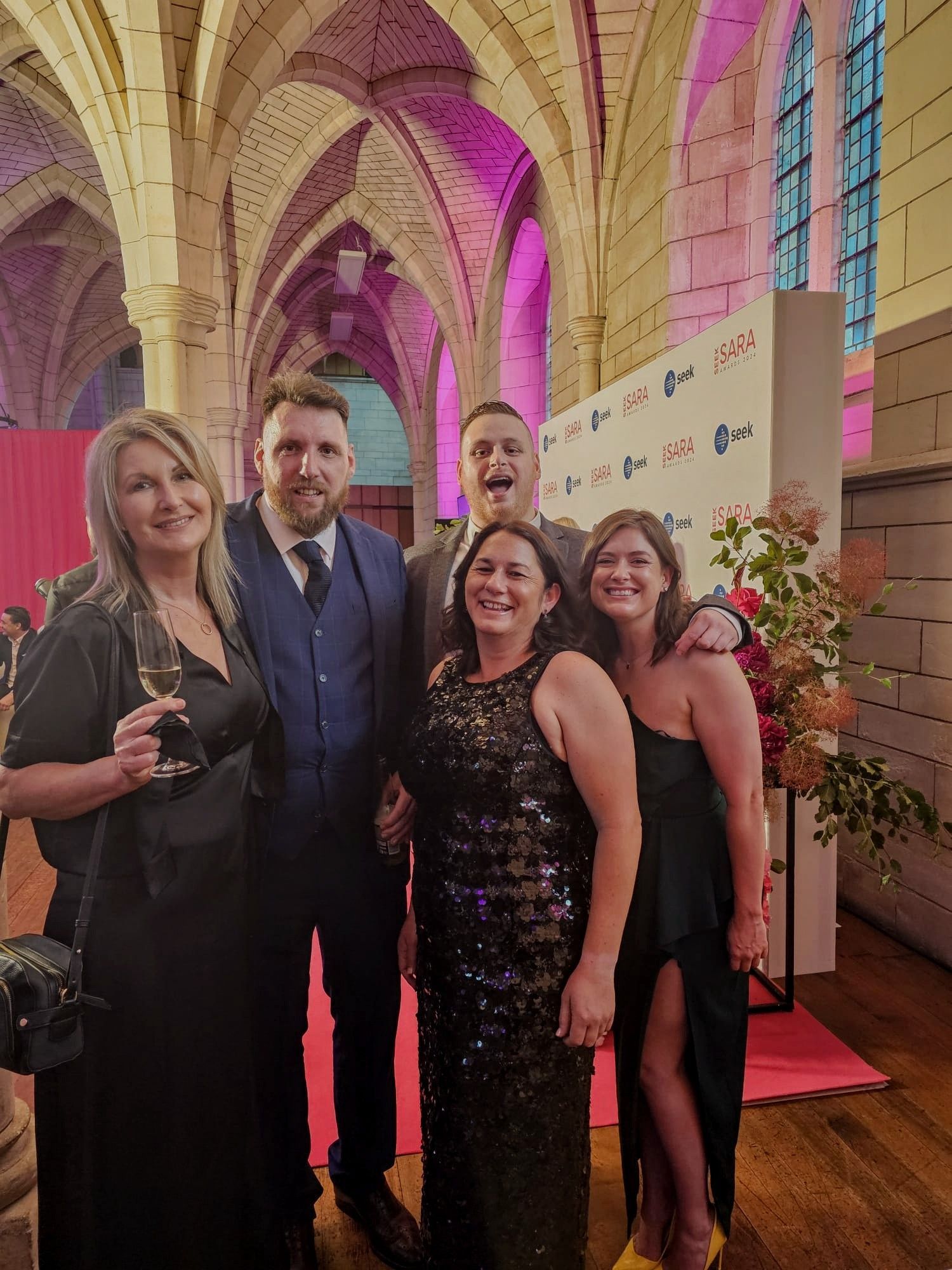 Five people posing at an event. A woman in a black sequined dress is centered. Pink lighting and ornate architecture.