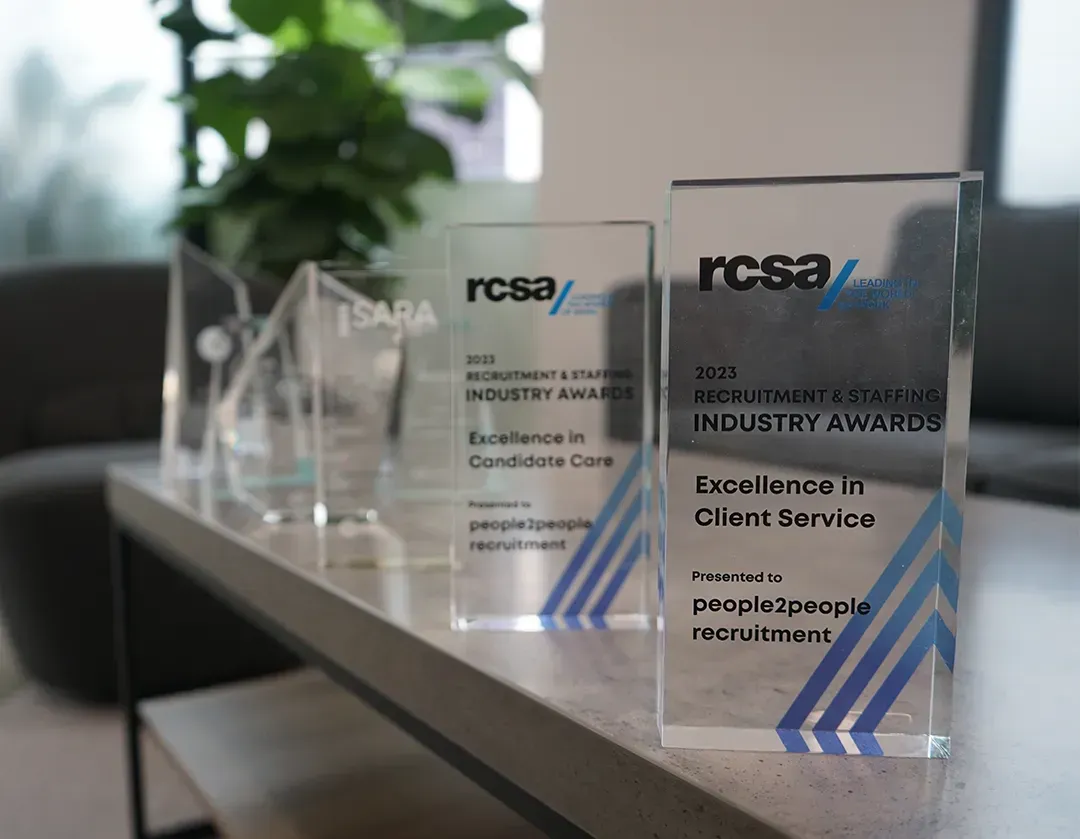 Several clear glass awards with blue accents on a table, plant and gray chair in background.