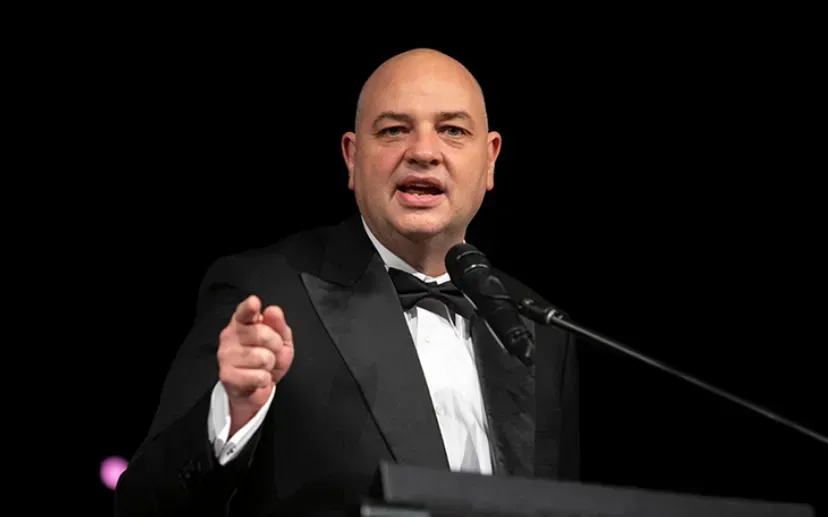 Man in tuxedo speaking at a podium, pointing, dark background.
