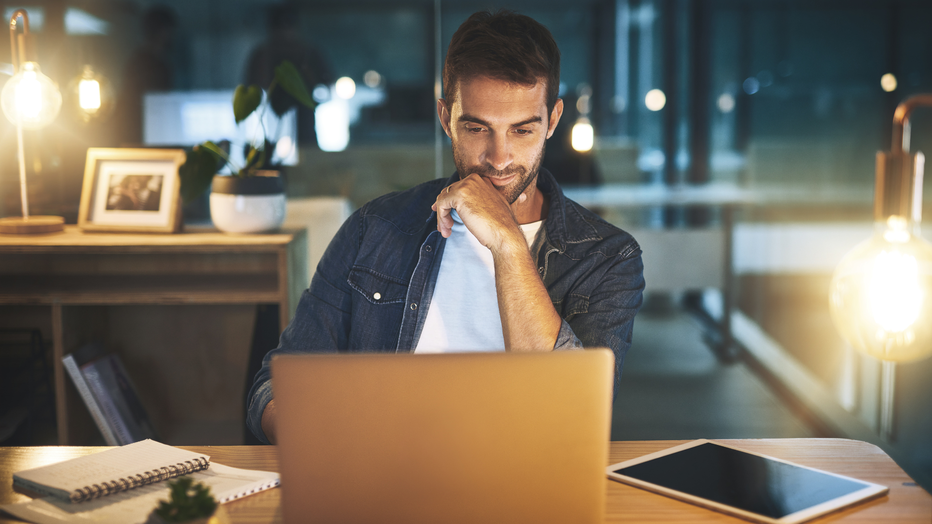 Man in denim shirt intently looks at laptop screen, hand on chin, at desk in dimly lit room.