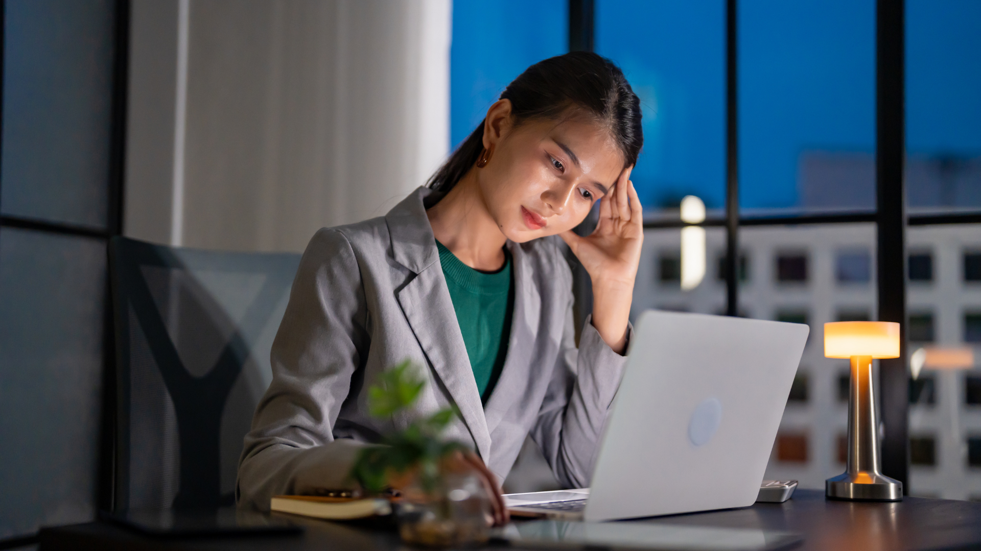 Woman in blazer at laptop, looks tired, hand on her forehead, at desk in office, night view.