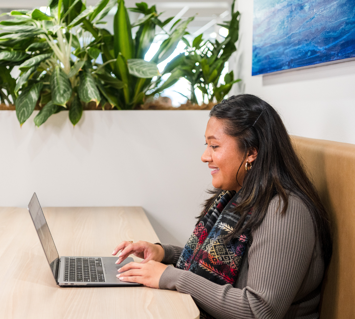 Woman smiles, typing on laptop at a light wood table, with plants in the background.