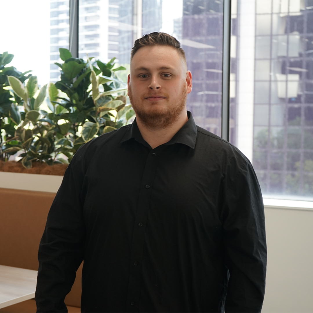 Man in a black shirt stands in an office setting, with a plant and a window with a cityscape visible.
