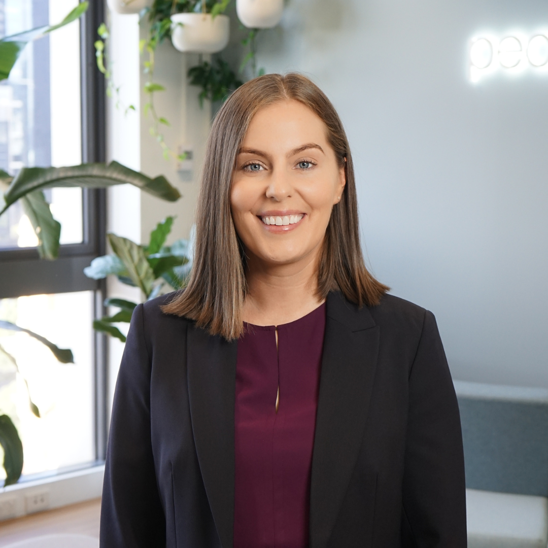 Woman in a dark blazer smiles at the camera, indoors with plants and a window.