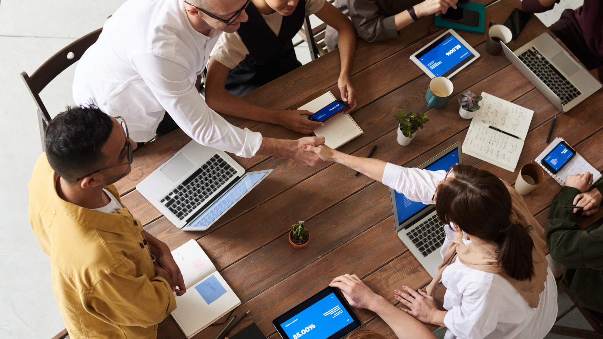 People shaking hands around a wooden table with laptops and tablets.