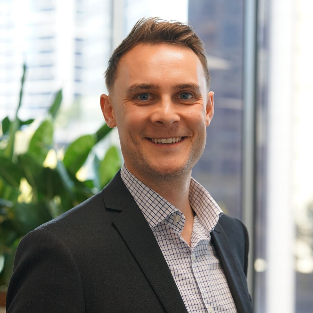 Man in a suit smiles near a window, office setting, collared shirt visible.