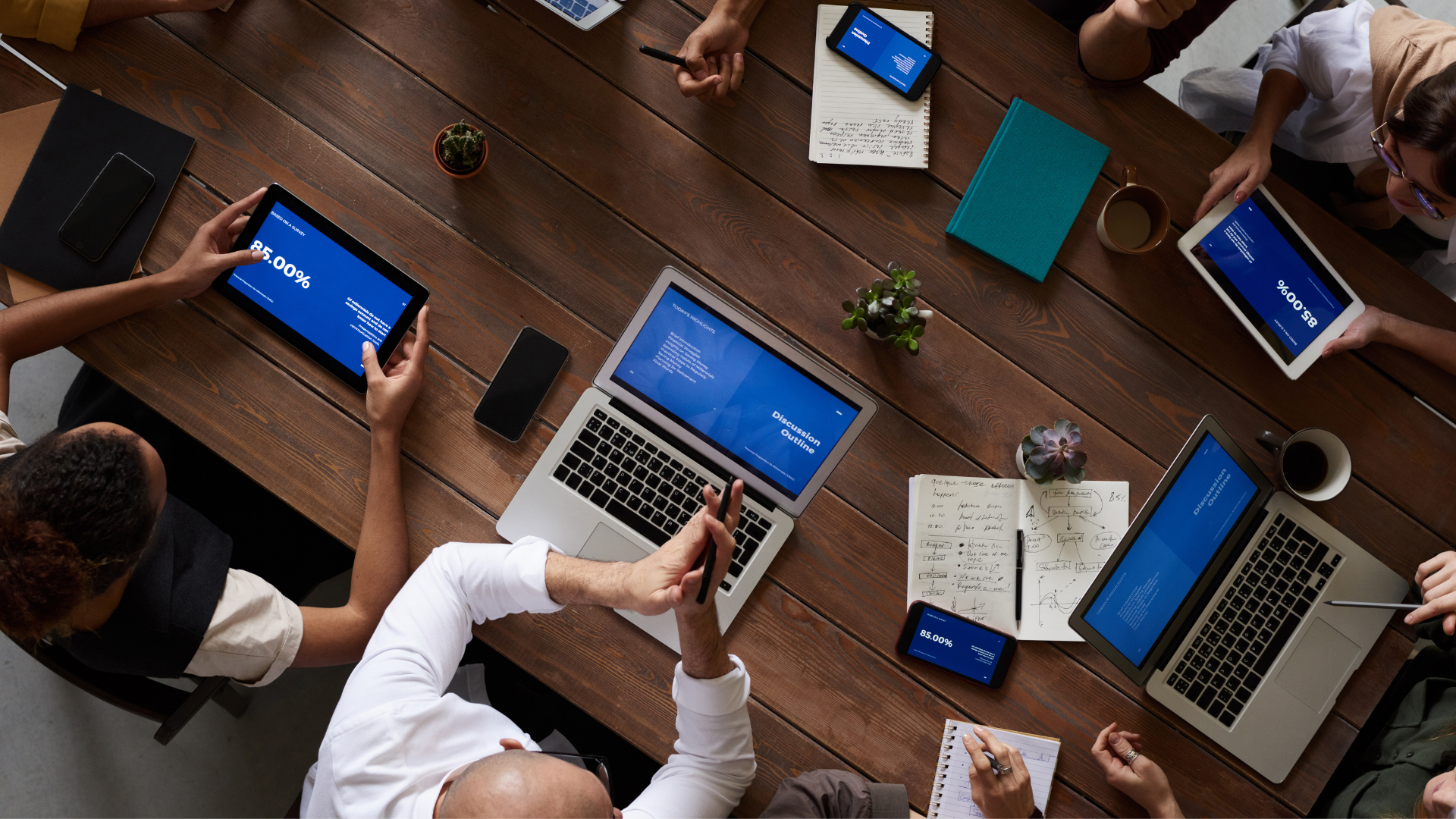 Group of people at a wooden table, laptops and tablets showing charts, discussing, overhead shot.