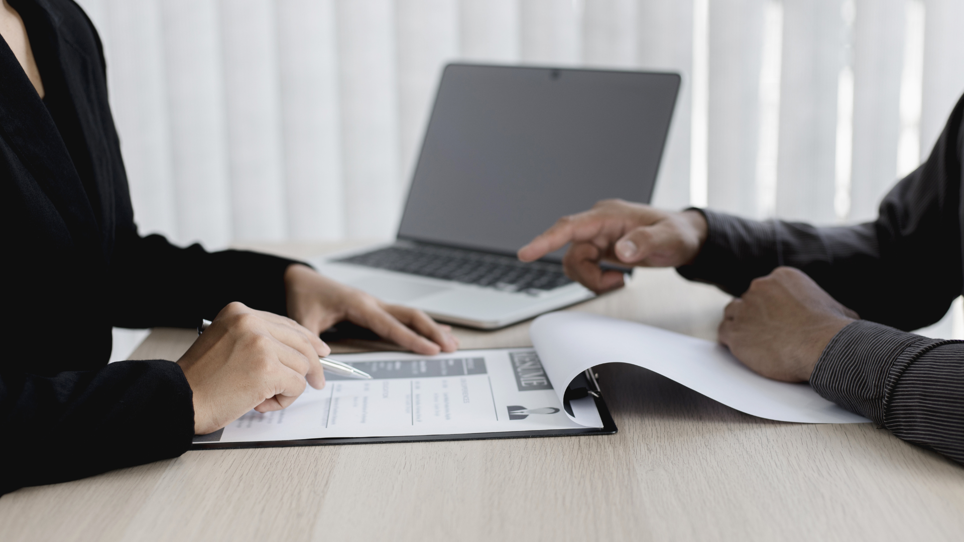 Two people at a desk review a document. One points; the other holds a pen near a resume. A laptop sits open.