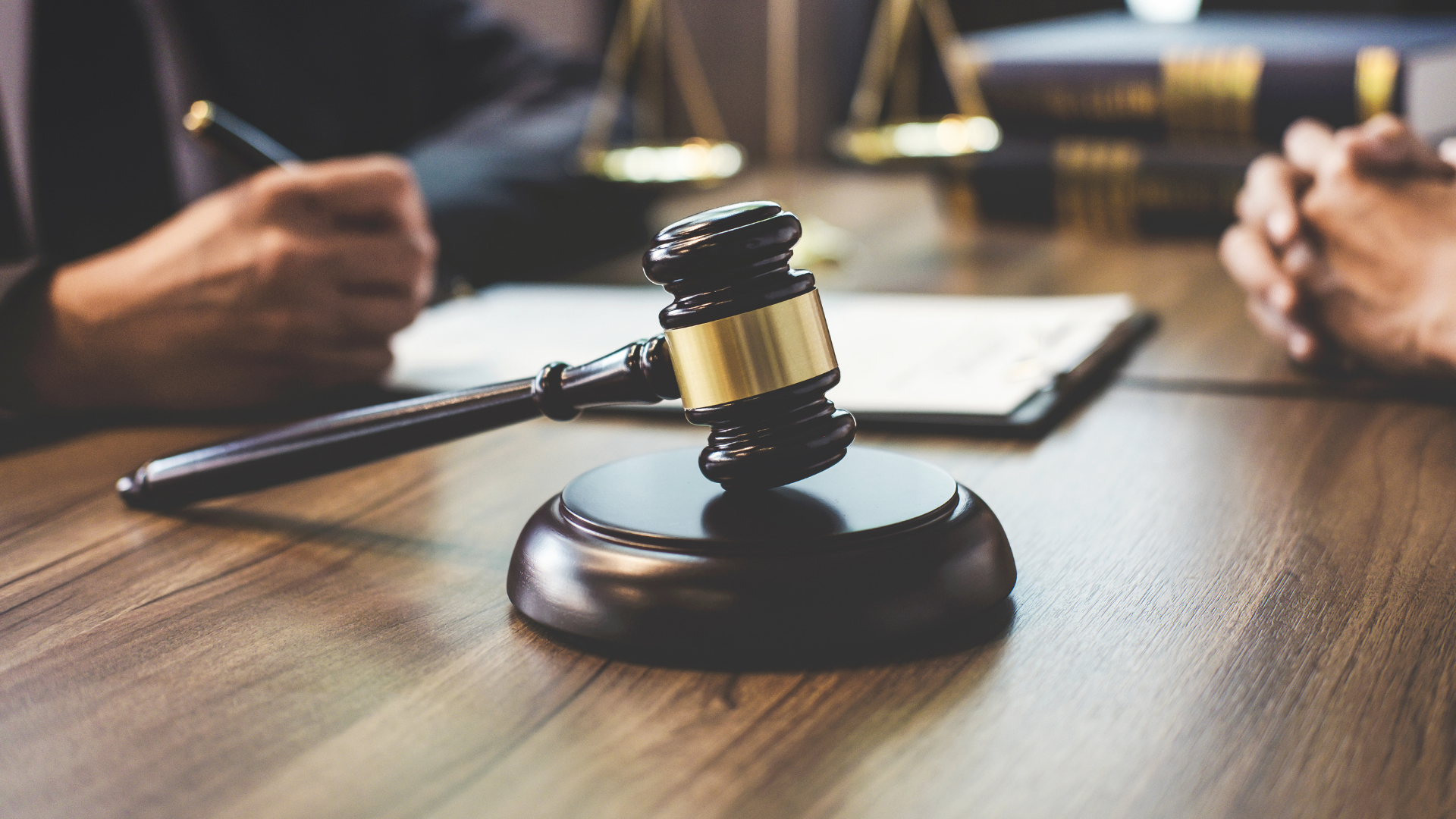 Gavel on desk, scales of justice and legal documents in background, two people.