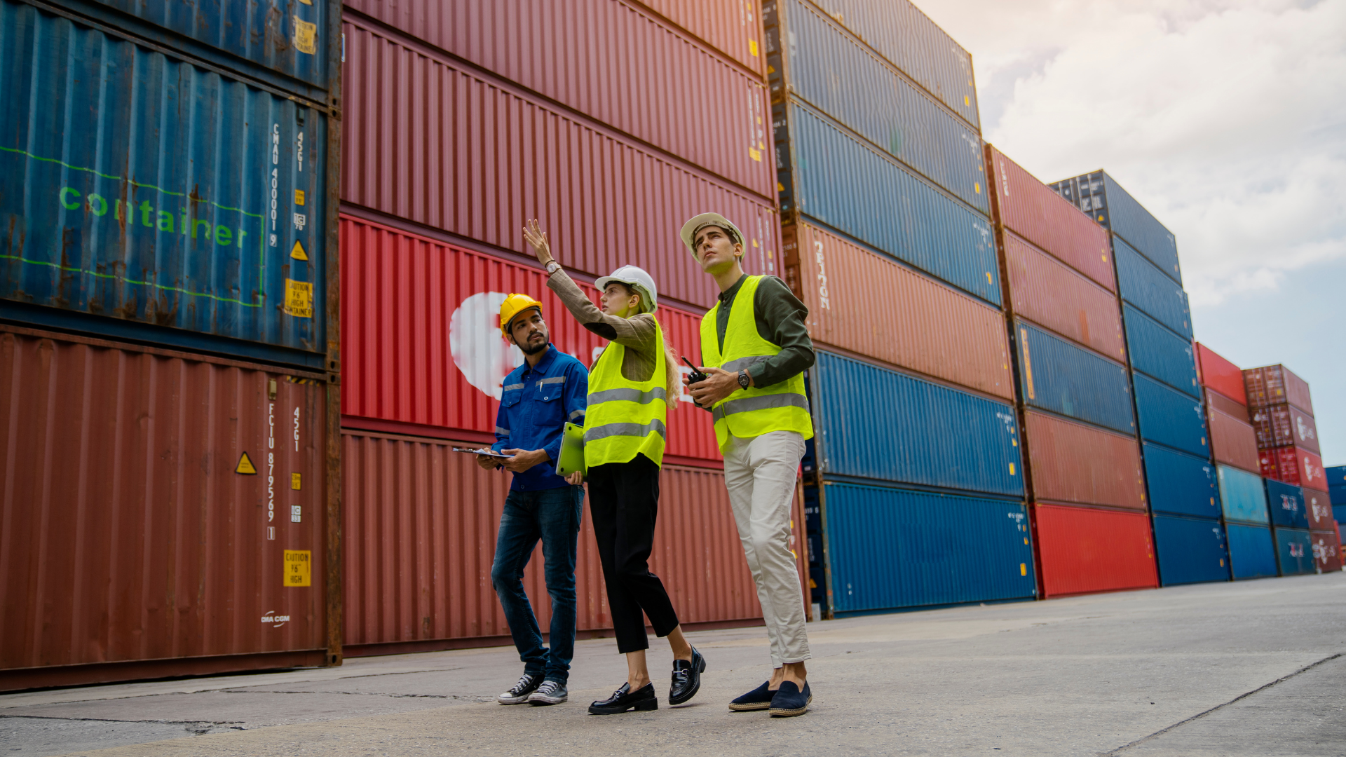 Three people inspecting shipping containers in a port. Two wear vests. One gestures upwards.