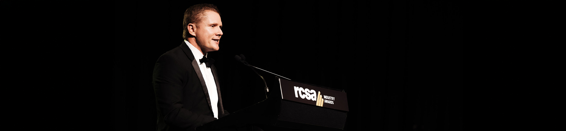 Person in a tuxedo speaking at a podium, set against a dark background.