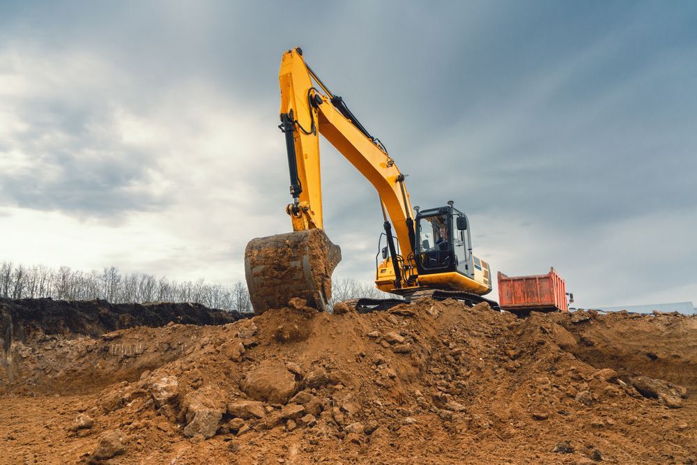 Yellow Excavator Digging Into a Brown Mound of Earth Under a Cloudy Sky — All Around Concreting in Lennox Head, NSW