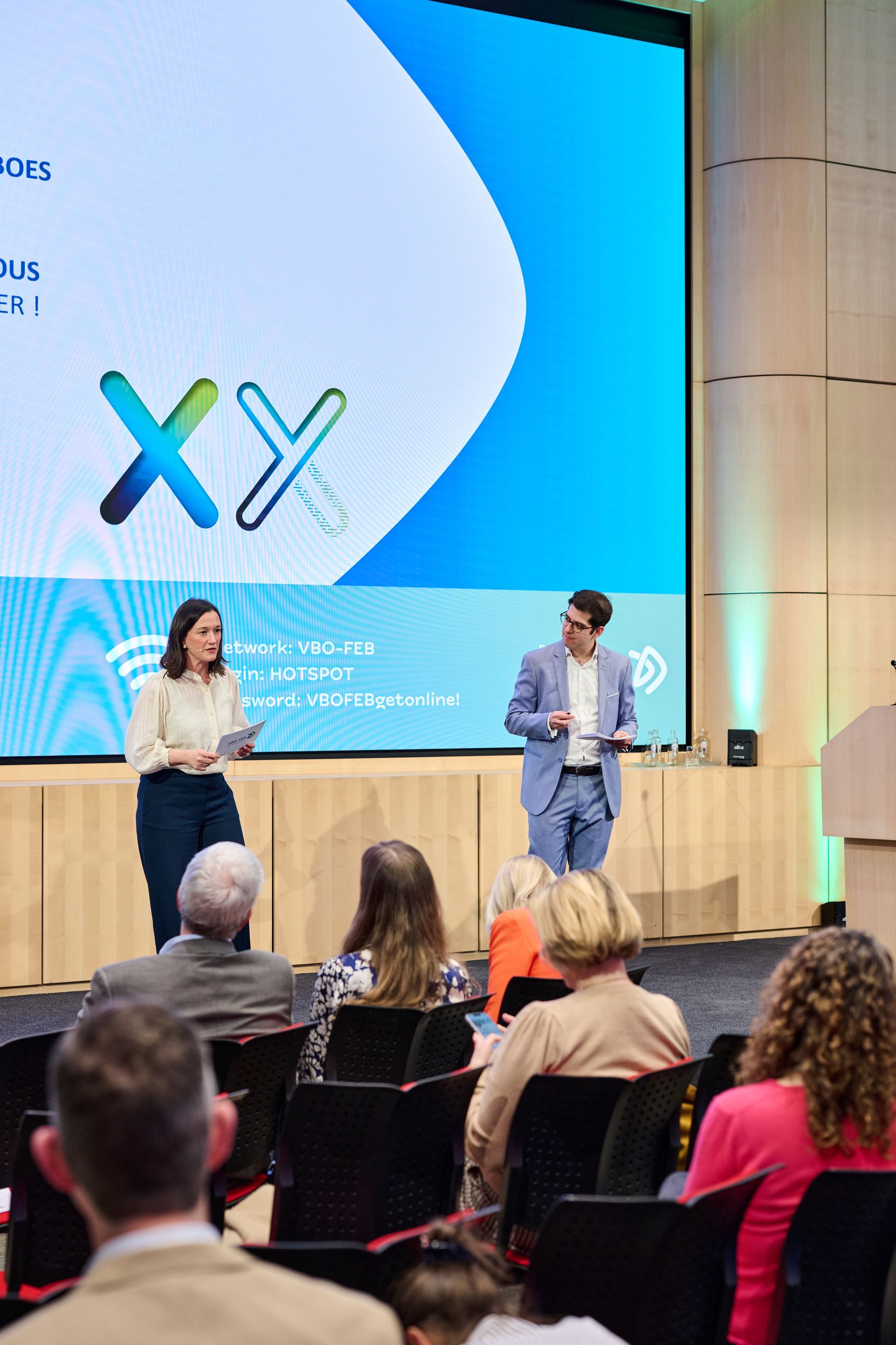 Two presenters on stage at a conference, speaking before an audience with a large blue screen backdrop.