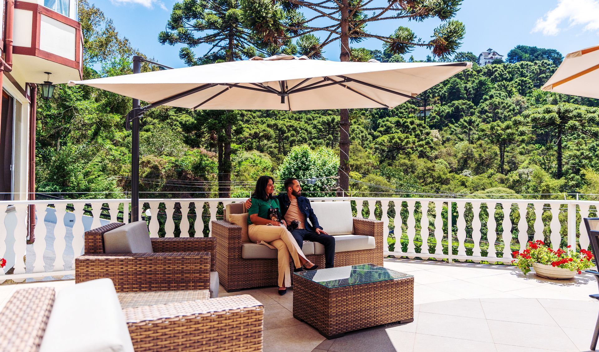 Duas pessoas estão sentadas em móveis de jardim sob um grande guarda-sol em uma varanda com vista para uma área arborizada.