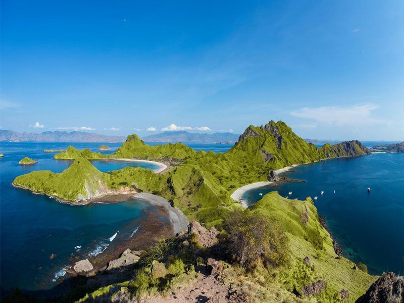 Vista panorâmica de colinas verdejantes e vibrantes, rodeadas por águas turquesa na Ilha de Padar, Indonésia, sob um céu limpo.