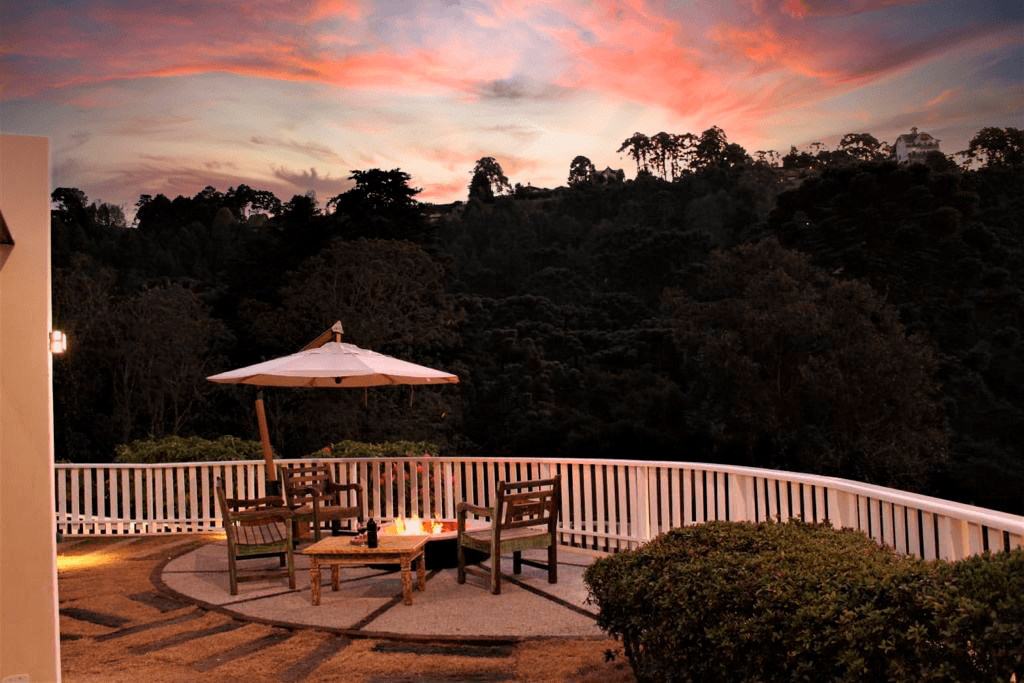 Um pátio para apreciar o pôr do sol com uma lareira externa, cadeiras de madeira, um guarda-sol e uma cerca branca com vista para uma encosta escura e arborizada.