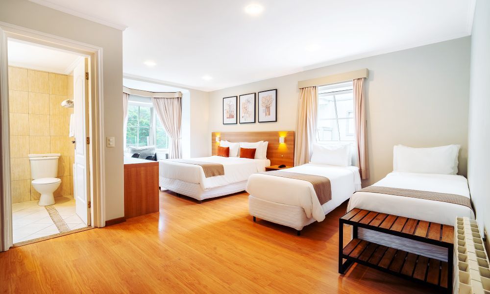 Um quarto de hotel espaçoso com duas camas, piso de madeira clara e um pequeno banheiro visível através de uma porta aberta.