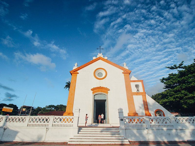 Uma igreja branca em estilo colonial com detalhes em laranja, uma cruz e um pequeno grupo de pessoas entrando sob um céu azul e nublado.