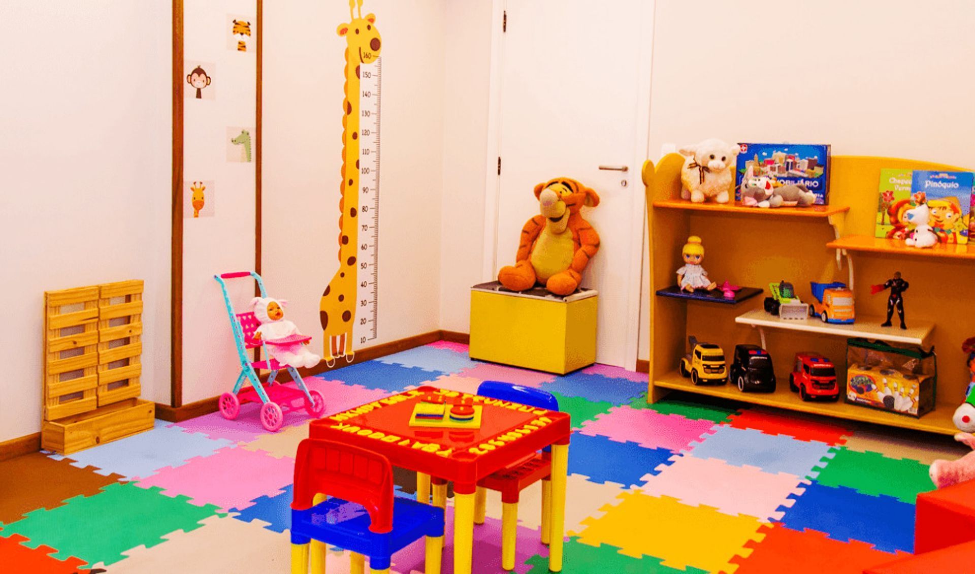 Uma sala de jogos infantil colorida apresenta uma pequena mesa e cadeiras, prateleiras para brinquedos, um carrinho de bebê de brinquedo e uma régua de altura em forma de girafa.
