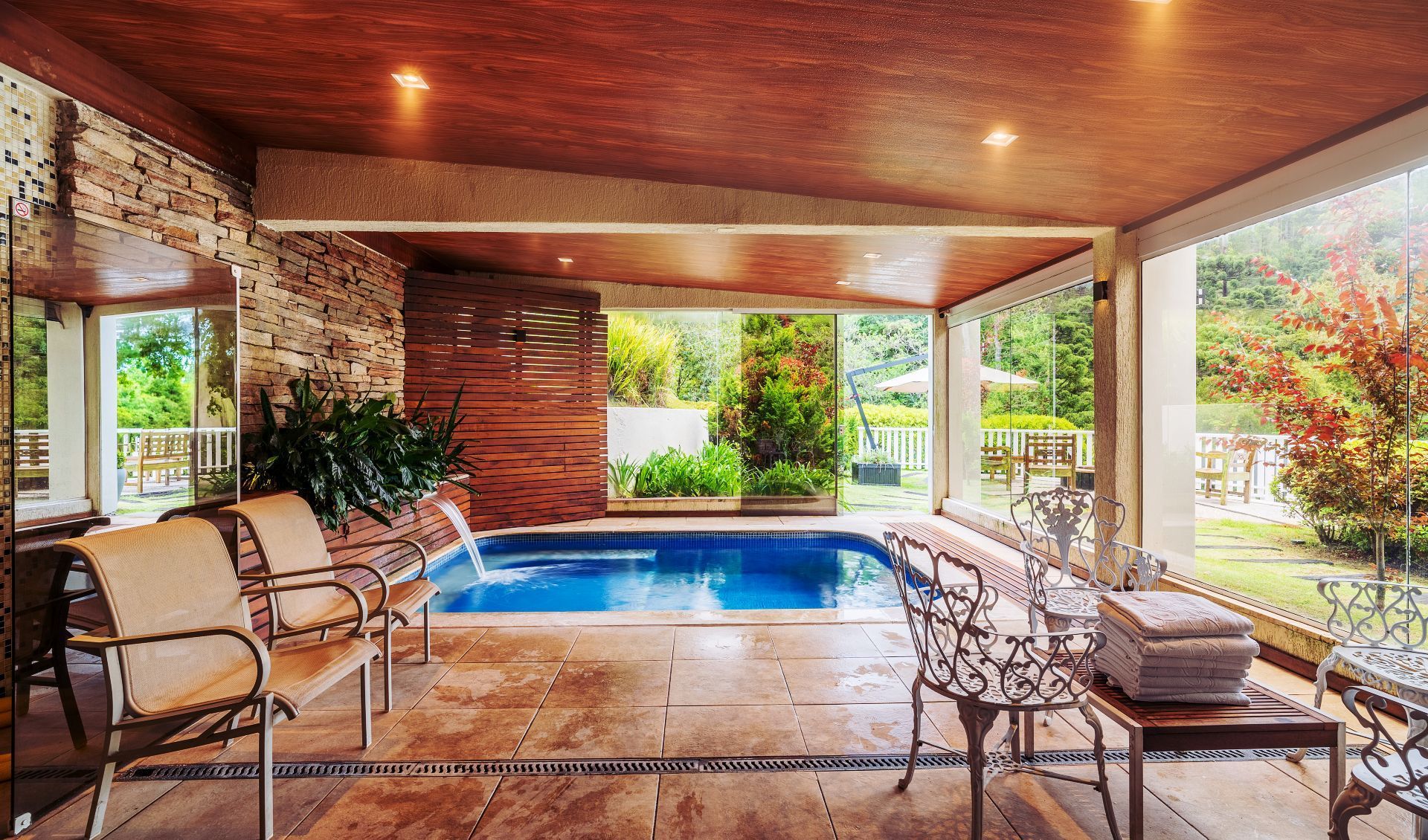 Uma área de piscina interna de luxo com piso de azulejo, paredes com detalhes em pedra, assentos confortáveis ​​e janelas do chão ao teto.
