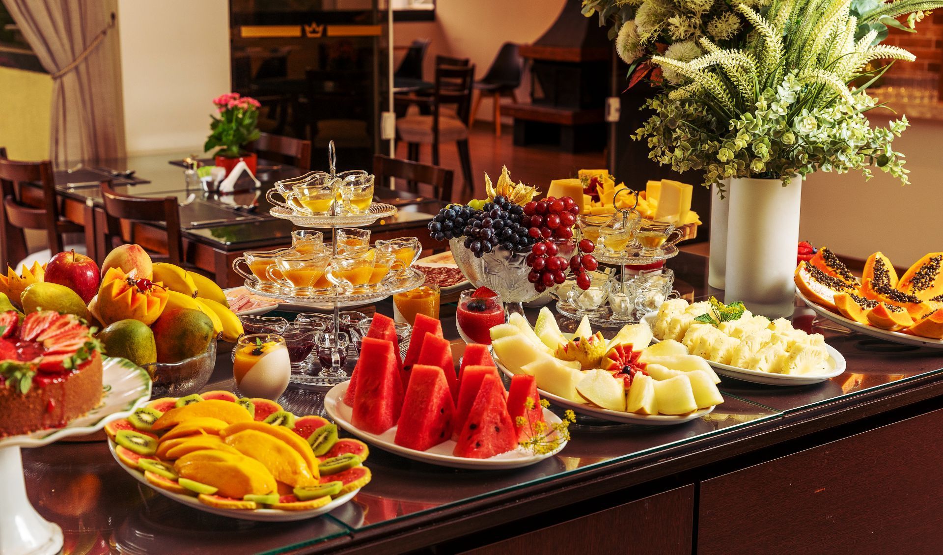 Um farto buffet de café da manhã com uma variedade de frutas frescas, bolos e pratos de vidro, disposto em uma mesa em uma sala de jantar com iluminação suave.