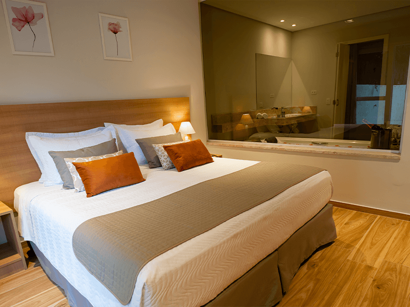 Um quarto de hotel com uma cama king-size com roupa de cama branca, cinza e laranja, e uma grande janela de vidro com vista para o banheiro.