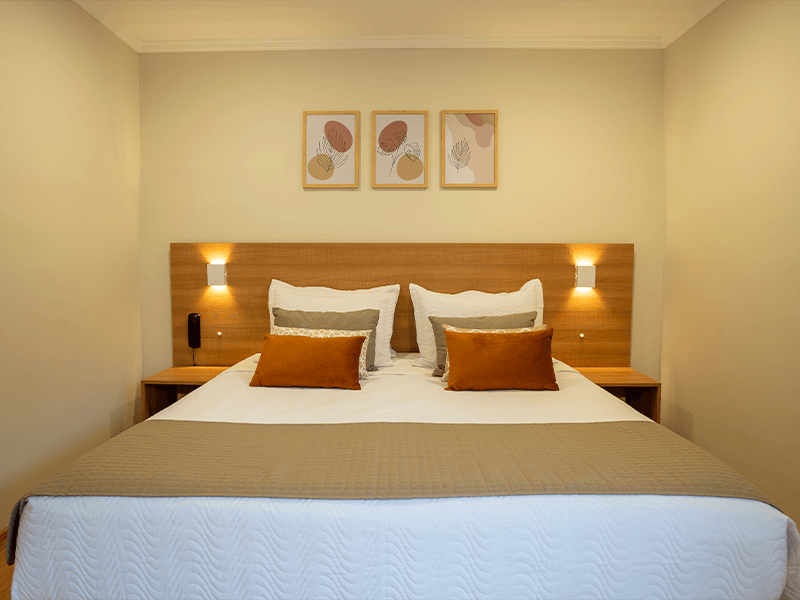 Um quarto de hotel moderno com cama queen-size, cabeceira de madeira, iluminação ambiente aconchegante e três gravuras abstratas na parede.