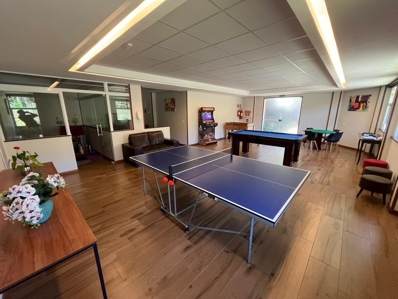 Uma sala de jogos com mesa de pingue-pongue, mesa de bilhar, mesa de cartas e uma pequena máquina de jogos eletrônicos sobre piso de madeira.