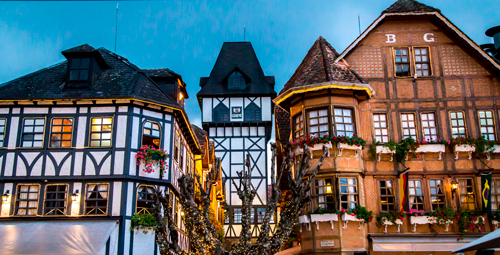 Uma cena de rua com três edifícios de vários andares, com estrutura de madeira e estilos arquitetônicos europeus, sob um céu azul.