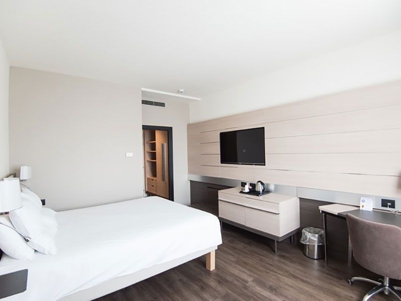 Um quarto de hotel moderno com cama arrumada, TV de tela plana em uma parede com painéis, mesa de apoio, escrivaninha e cadeira de escritório.