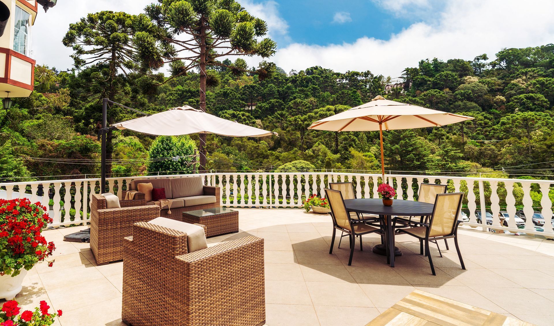 Um terraço ensolarado ao ar livre com cadeiras de vime, uma mesa de jantar sob dois guarda-sóis e vista para uma exuberante floresta verde.