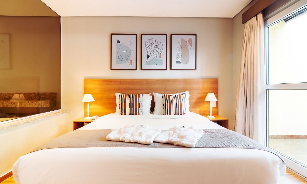 Um quarto de hotel moderno com uma cama branca impecavelmente arrumada, duas almofadas estampadas, candeeiros de cabeceira e obras de arte emolduradas na parede.