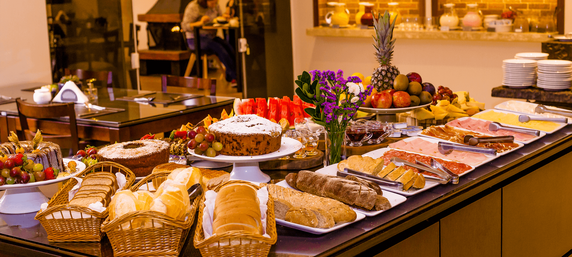 Um buffet de café da manhã com cestas de pão, bolos e uma travessa de frutas com um abacaxi em uma longa mesa de serviço.