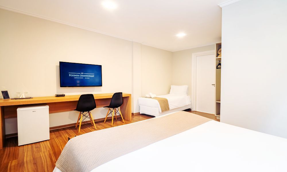 Um quarto de hotel moderno com uma escrivaninha de madeira, duas cadeiras pretas, uma TV, um frigobar e vista para duas camas com lençóis brancos.